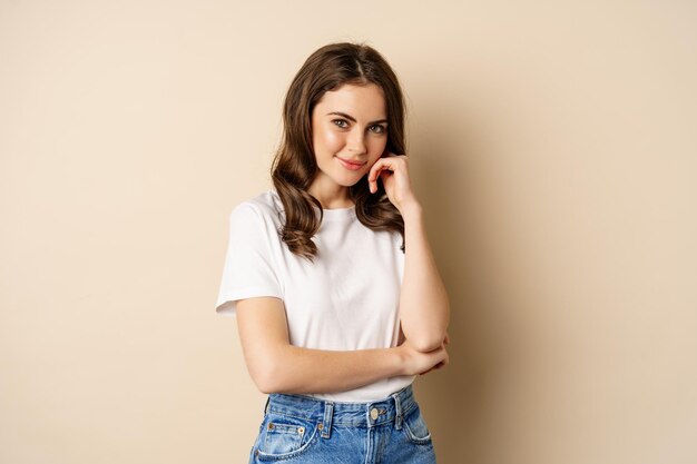 Retrato de una hermosa joven con camiseta blanca, sonriendo y luciendo feliz, posando sobre un fondo beige.