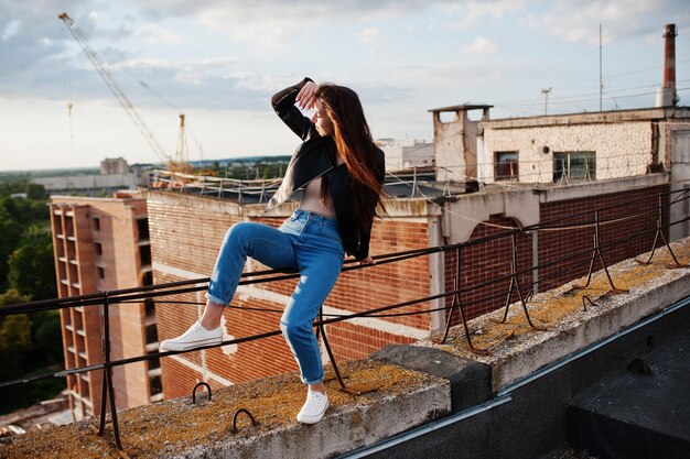 Retrato de una hermosa joven con chaqueta de cuero negra, jeans y zapatillas sentadas en pasamanos en el techo con una vista pintoresca de un parque