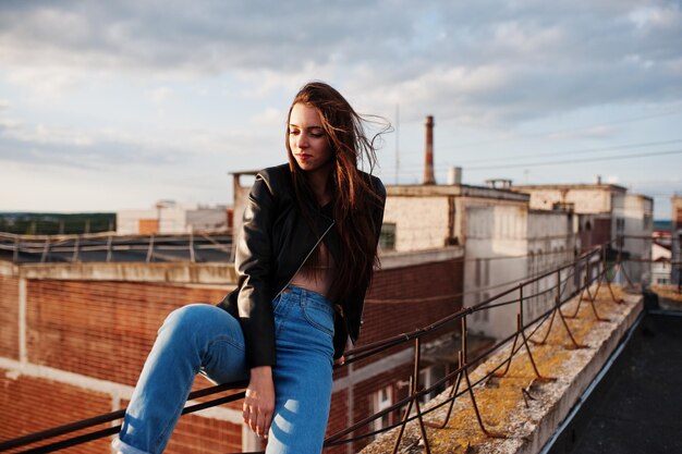 Retrato de una hermosa joven con chaqueta de cuero negra, jeans y zapatillas sentadas en pasamanos en el techo con una vista pintoresca de un parque