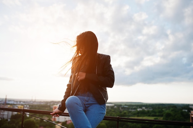 Retrato de una hermosa joven con chaqueta de cuero negra, jeans y zapatillas sentadas en pasamanos en el techo con una vista pintoresca de un parque