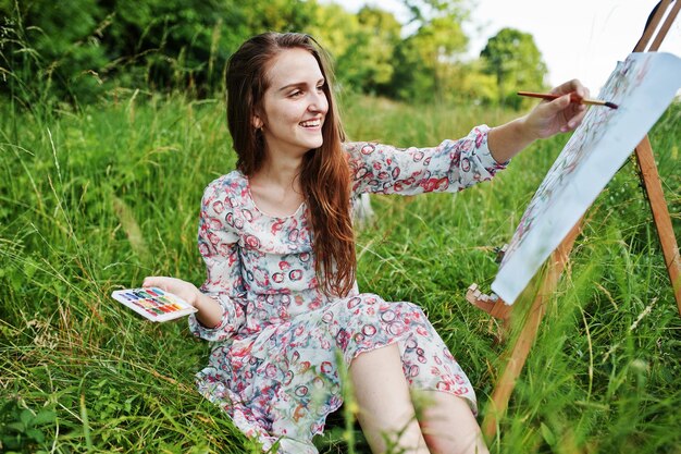 Retrato de una hermosa joven feliz con un hermoso vestido sentada en el césped y pintando en papel con acuarelas