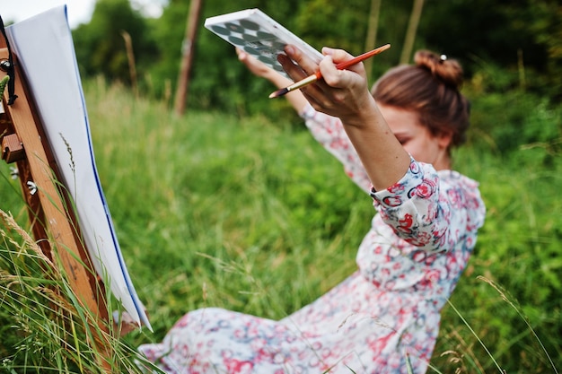 Retrato de una hermosa joven feliz con un hermoso vestido sentada en el césped y pintando en papel con acuarelas