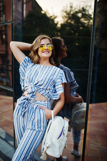 Foto gratuita retrato de una hermosa joven con gafas de sol amarillas y a rayas posando con una mochila de cuero blanco en un balcón de un moderno edificio de vidrio en la ciudad