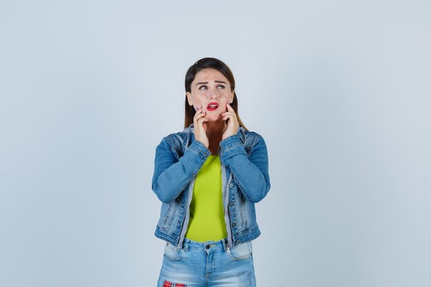 Retrato de hermosa joven manteniendo los dedos en las mejillas mientras mira hacia arriba en traje de mezclilla y mira pensativa vista frontal