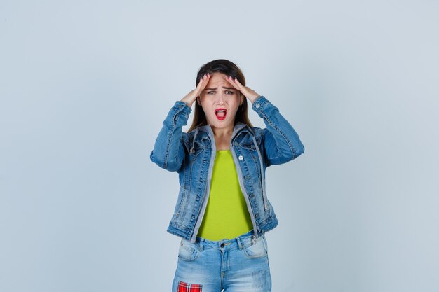 Retrato de hermosa joven manteniendo las manos sobre la cabeza en traje de mezclilla y mirando olvidadizo vista frontal