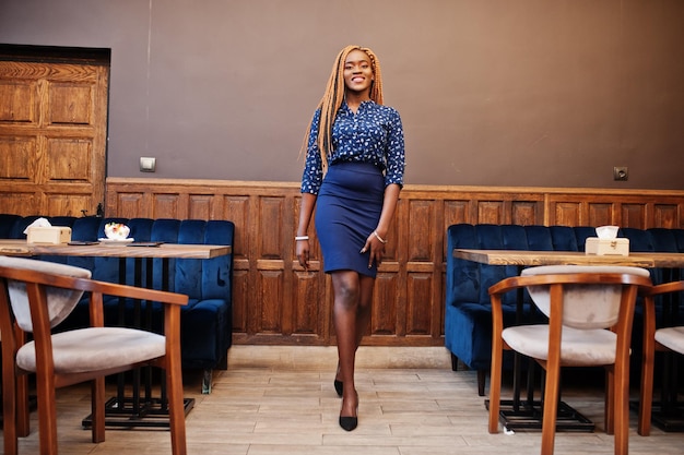 Retrato de una hermosa joven mujer de negocios africana con rastas en blusa azul y falda posada en un café