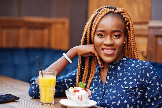 Retrato de una hermosa joven mujer de negocios africana con rastas en blusa azul y falda sentada en un café con helado y jugo de piña