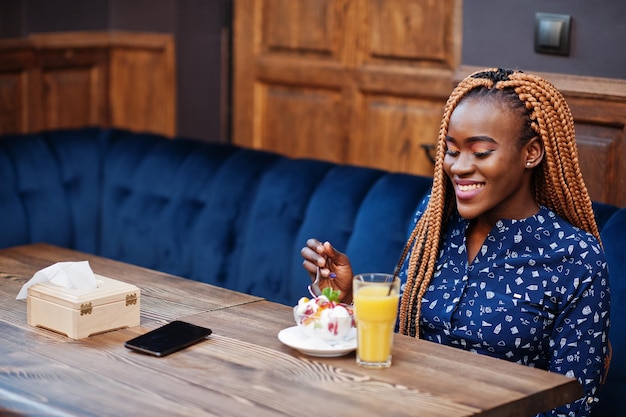 Retrato de una hermosa joven mujer de negocios africana con rastas en blusa azul y falda sentada en un café con helado y jugo de piña