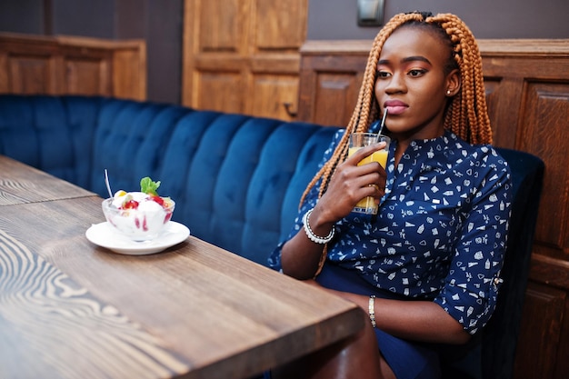 Retrato de una hermosa joven mujer de negocios africana con rastas en blusa azul y falda sentada en un café con helado y jugo de piña