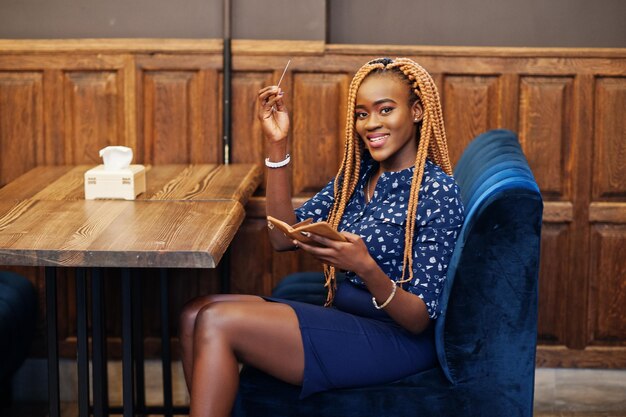 Retrato de una hermosa joven mujer de negocios africana vestida con blusa azul y falda sentada en el restaurante y con tarjeta de crédito en la mano