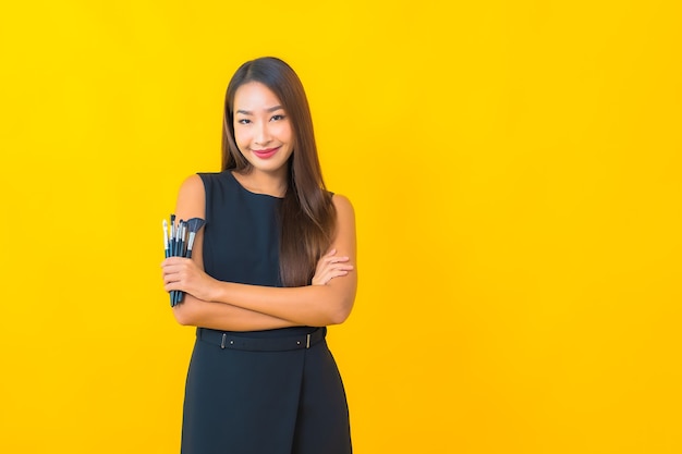 Retrato hermosa joven mujer de negocios asiática con maquillaje cepillo cosmético sobre fondo amarillo