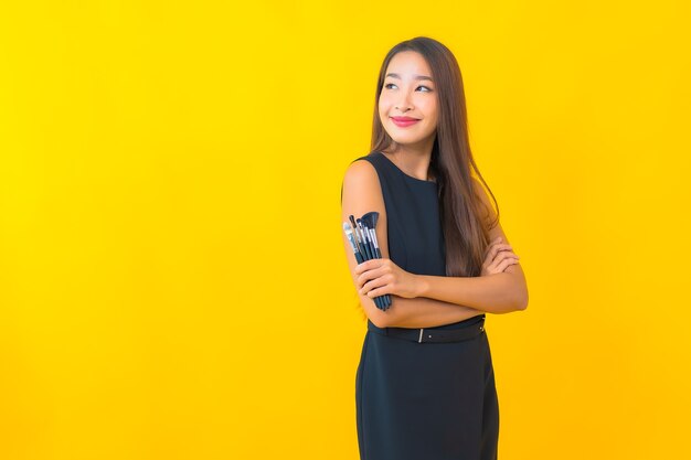 Retrato hermosa joven mujer de negocios asiática con maquillaje cepillo cosmético sobre fondo amarillo