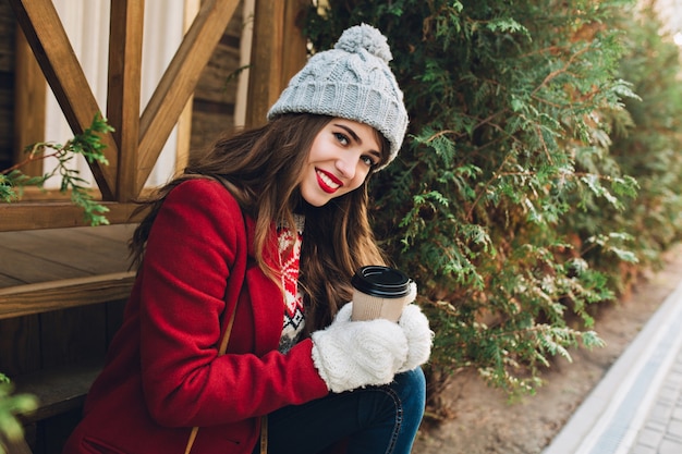 Retrato hermosa joven con pelo largo en abrigo rojo sentado en escaleras de madera al aire libre. Ella tiene gorro de punto gris, guantes blancos, toma café y sonríe.