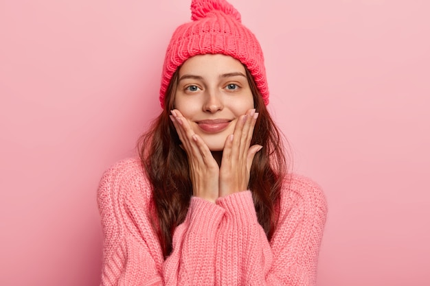 El retrato de una hermosa joven sonríe agradablemente, mantiene ambas palmas en las mejillas, mira con alegría a la cámara, tiene un aspecto relajado, usa un suéter y gorro de invierno de punto, modelos de interior sobre una pared de estudio rosa