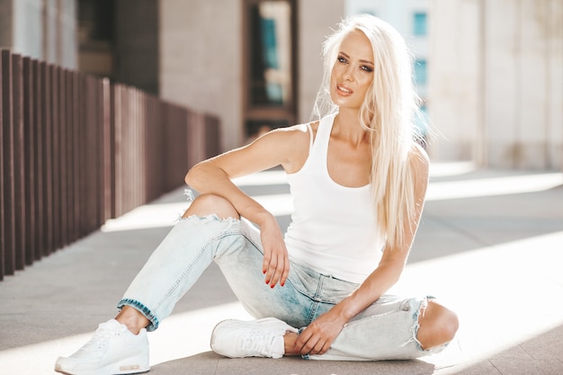 Retrato de hermosa linda chica rubia en camiseta blanca y jeans posando al aire libre. Linda chica sentada en el asfalto en la calle