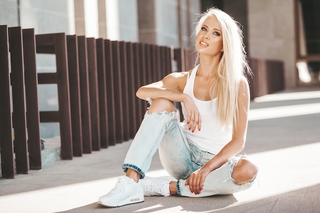 Retrato de hermosa linda chica rubia en camiseta blanca y jeans posando al aire libre. Linda chica sentada en el asfalto en la calle