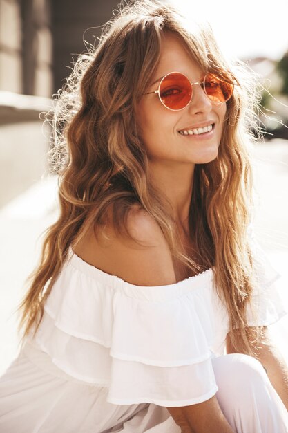 Retrato de la hermosa modelo de adolescente rubia linda sin maquillaje en ropa de vestido blanco hipster de verano sentado en el fondo de la calle