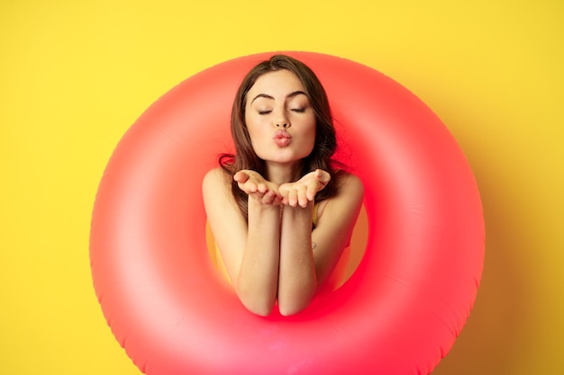 Retrato de una hermosa modelo femenina posando con un anillo de playa de natación rosa, enviando un beso al aire, mwah coqueta, de pie sobre un fondo amarillo