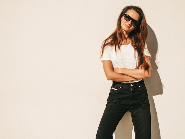 Foto gratuita retrato de hermosa modelo en gafas de sol. mujer vestida con jeans y camiseta blanca de verano hipster. chica de moda posando junto a la pared en la calle