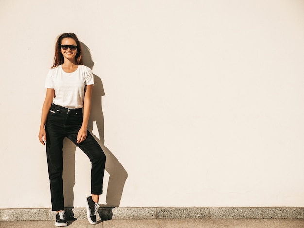 Retrato de hermosa modelo en gafas de sol. Mujer vestida con jeans y camiseta blanca de verano hipster. Mujer de moda posando junto a la pared en la calle