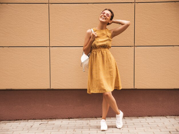 Retrato de la hermosa modelo hipster sonriente vestido con vestido amarillo de verano. Chica de moda posando en la calle. Mujer divertida y positiva divirtiéndose