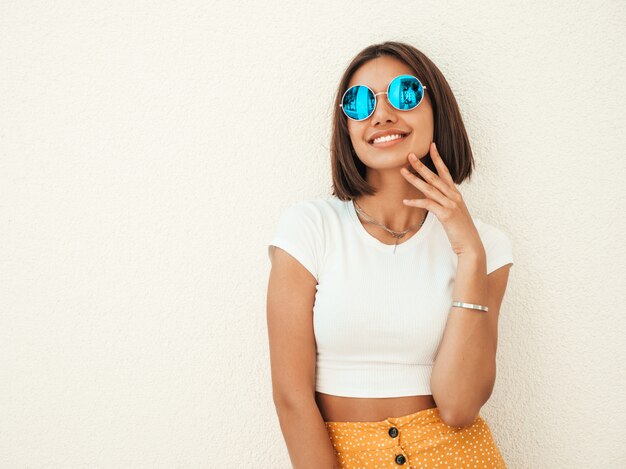 Retrato de la hermosa modelo morena sonriente vestida con ropa hipster de verano. Chica de moda posando en la calle cerca de la pared blanca. Mujer divertida y positiva divirtiéndose