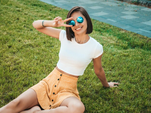 Retrato de la hermosa modelo morena sonriente vestida con ropa hipster de verano. Moda niña sentada en el césped en el parque. Mujer divertida y positiva divirtiéndose. Muestra el signo de la paz