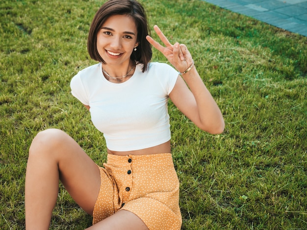 Retrato de la hermosa modelo morena sonriente vestida con ropa hipster de verano. Moda niña sentada en el césped en el parque. Mujer divertida y positiva divirtiéndose. Muestra el signo de la paz