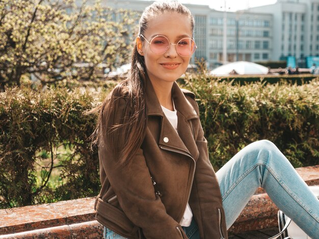 Retrato de la hermosa modelo morena sonriente vestida con ropa de jeans y chaqueta hipster de verano Chica de moda sentada en el banco en la calle Mujer divertida y positiva en gafas de sol