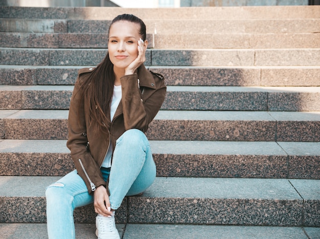 Retrato de la hermosa modelo morena vestida con ropa de jeans y chaqueta hipster de verano. Moda niña sentada en pasos en el fondo de la calle. Mujer divertida y positiva