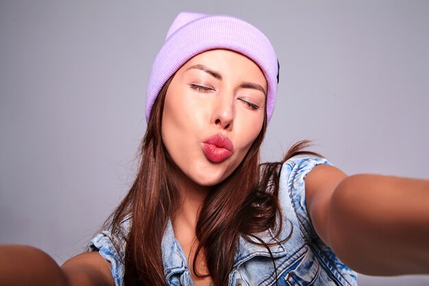 Retrato de la hermosa modelo de mujer morena linda en ropa casual de verano jeans sin maquillaje en gorro púrpura haciendo foto selfie en teléfono aislado en gris. Dando beso de aire