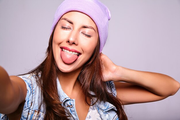 Retrato de la hermosa modelo de mujer morena linda en ropa casual de verano jeans sin maquillaje en gorro púrpura haciendo foto selfie en teléfono aislado en gris. Mostrando su lengua