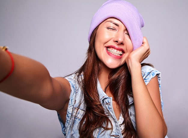 Retrato de la hermosa modelo de mujer morena linda sonriente en ropa casual de verano jeans sin maquillaje en gorro púrpura haciendo foto selfie en teléfono aislado en gris