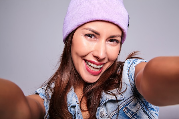 Retrato de la hermosa modelo de mujer morena linda sonriente en ropa casual de verano jeans sin maquillaje en gorro púrpura haciendo foto selfie en teléfono aislado en gris