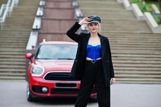 Retrato de una hermosa modelo rubia de moda sexy con gorra y corsé azul negro con maquillaje brillante cerca del coche rojo de la ciudad