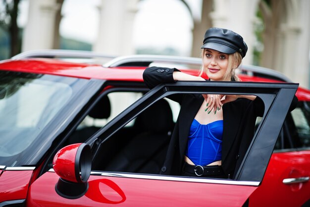 Retrato de una hermosa modelo rubia de moda sexy con gorra y corsé azul negro con maquillaje brillante cerca del coche rojo de la ciudad