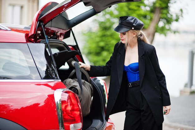 Retrato de una hermosa modelo rubia de moda sexy con gorra y todo negro con maquillaje brillante cerca del auto rojo de la ciudad puso su bolso en el baúl