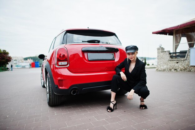 Retrato de una hermosa modelo rubia de moda sexy con gorra y todo negro con maquillaje brillante cerca del coche rojo de la ciudad