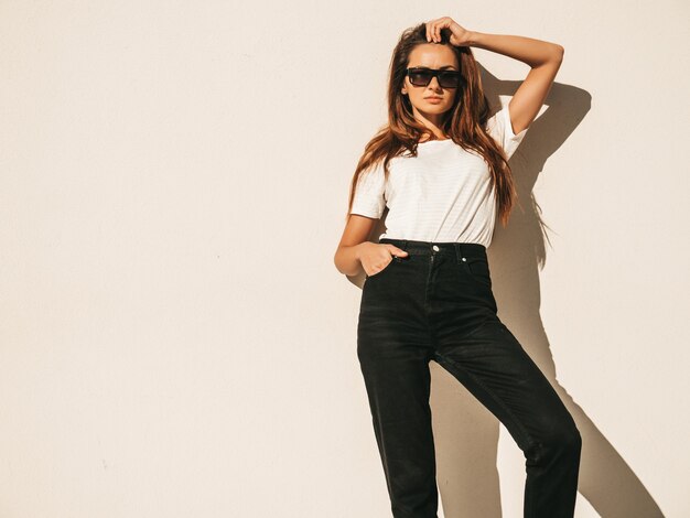 Retrato de hermosa modelo sonriente en gafas de sol. Mujer vestida con jeans y camiseta blanca hipster de verano. Posando junto a la pared en la calle