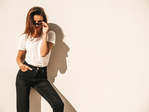 Retrato de hermosa modelo sonriente en gafas de sol. Mujer vestida con jeans y camiseta blanca hipster de verano. Posando junto a la pared en la calle