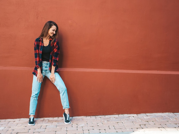 Retrato de hermosa modelo sonriente. Mujer vestida con jeans y camisa a cuadros de verano hipster. Chica de moda posando junto a la pared en la calle