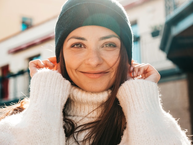 Retrato de hermosa modelo sonriente. Mujer vestida con suéter blanco cálido hipster y gorro. Ella posando en la calle