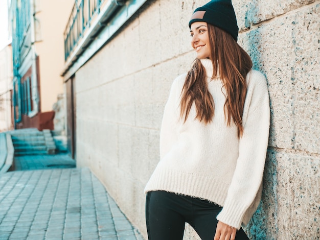 Retrato de hermosa modelo sonriente. Mujer vestida con suéter blanco cálido hipster y gorro. Posando en la calle