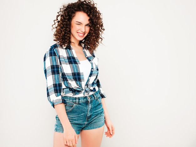 Retrato de la hermosa modelo sonriente con peinado afro rizos vestido con ropa hipster de verano. Chica despreocupada sexy posando en estudio sobre fondo gris. Moda mujer divertida y positiva guiño