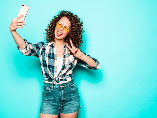 Retrato de hermosa modelo sonriente con peinado afro rizos vestido con ropa hipster de verano. Chica despreocupada sexy posando en estudio sobre fondo gris. Mujer divertida de moda toma selfie foto