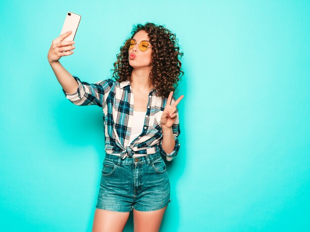 Retrato de hermosa modelo sonriente con peinado afro rizos vestido con ropa hipster de verano. Chica despreocupada sexy posando en estudio sobre fondo gris. Mujer divertida de moda toma selfie foto