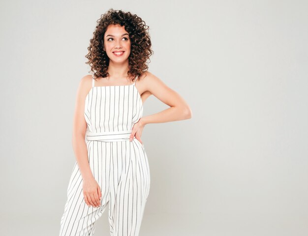 Retrato de hermosa modelo sonriente con peinado afro rizos vestido con ropa hipster de verano. Chica despreocupada sexy posando en estudio sobre fondo gris. Mujer divertida y positiva de moda