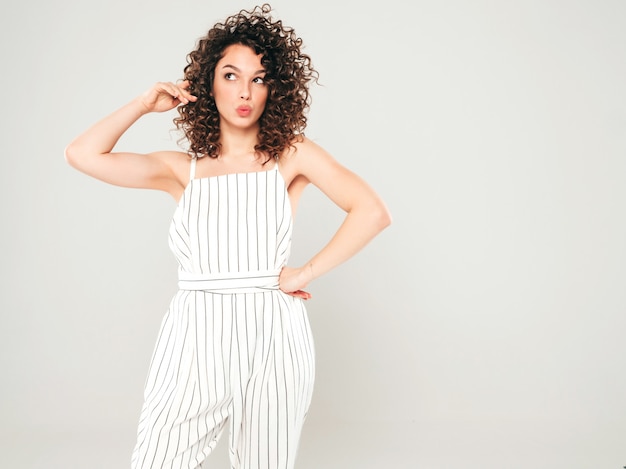 Retrato de hermosa modelo sonriente con peinado afro rizos vestido con ropa hipster de verano. Chica despreocupada sexy posando en estudio sobre fondo gris. Mujer divertida y positiva de moda