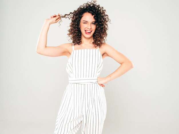 Retrato de hermosa modelo sonriente con peinado afro rizos vestido con ropa hipster de verano. Chica despreocupada sexy posando en estudio sobre fondo gris. Mujer divertida y positiva de moda