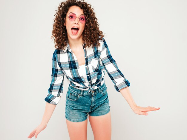 Retrato de hermosa modelo sonriente con peinado afro rizos vestido con ropa hipster de verano. Chica despreocupada sexy posando en estudio sobre fondo gris. Mujer divertida y positiva de moda